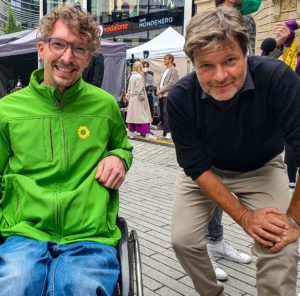 Dennis und Robert Habeck blicken gemeinsam lächelnd in die Kamera. Dennis trägt eine grüne Sportjacke von B90/Die Grünen. Robert trägt ein schwarzes Hemd. Im Hintergrund ist ein Wochenmarkt zu sehen.