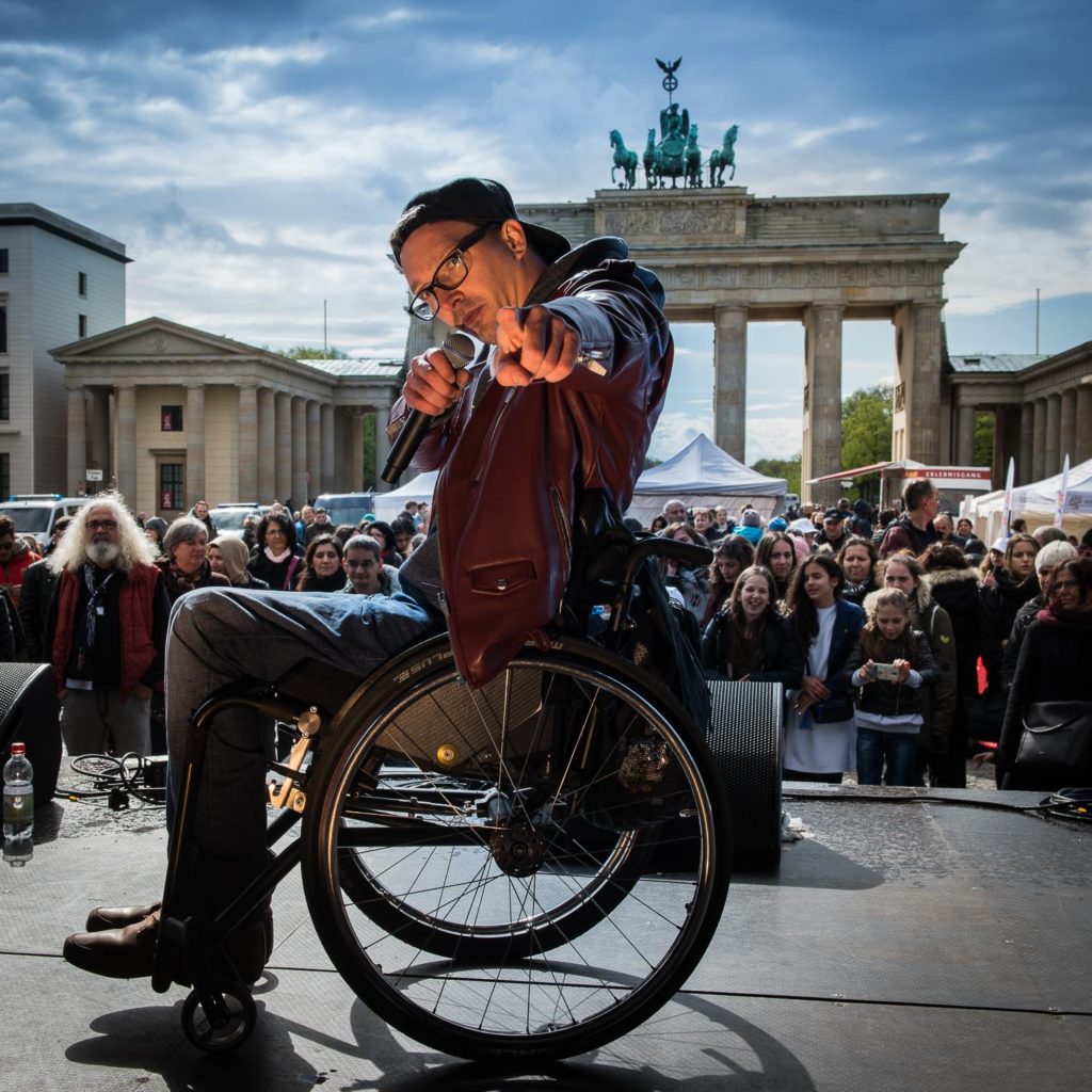 Dennis befindet sich vor dem Brandenburger-Tor. Er trägt eine Capy nach hinten gerichtet. Mit einem Mikro in der einen Hand, zeigt er mit der anderen Hand weisend in die Kamera. Er performt einen seiner Songs. Zwischen ihm und den Brandenburger-Tor ist eine große Menschenmasse, die ihm begeistert zuhört.