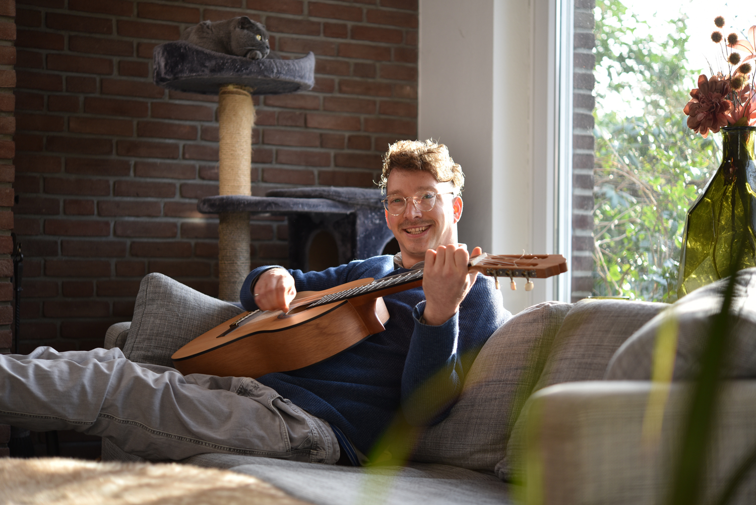 Dennis sitzt in seinem Wohnzimmer auf dem Sofa. Er hält seine Gitarre in der Hand und spielt diese. Im Hintergrund sieht man durch das Fenster einen Ausschnitt seines Gartens.