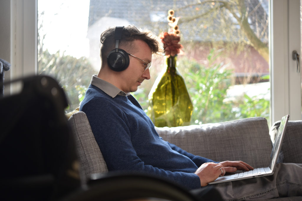 Dennis hat Kopfhörer auf und liegt auf der Couch. Er arbeitet an seinen Laptop. Hinter Dennis ist ein großes Fenster. Die Sonne scheint durch das Fenster.