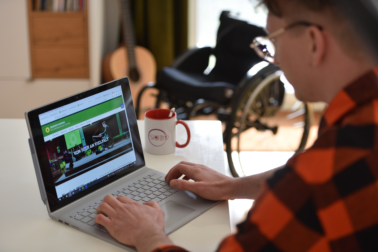 Dennis Sonne sitzt an seinem Notebook. Er trägt ein Holzfällerhemd (Rot-Schwarz-Kariert). Auf dem Bildschirm seines Notebooks ist zu sehen, wir er seine Homepage bearbeitet. Neben dem Notebook befindet sich eine Tasse. Im Hintergrund sieht man seinen Rollstuhl und seine Gitarre.