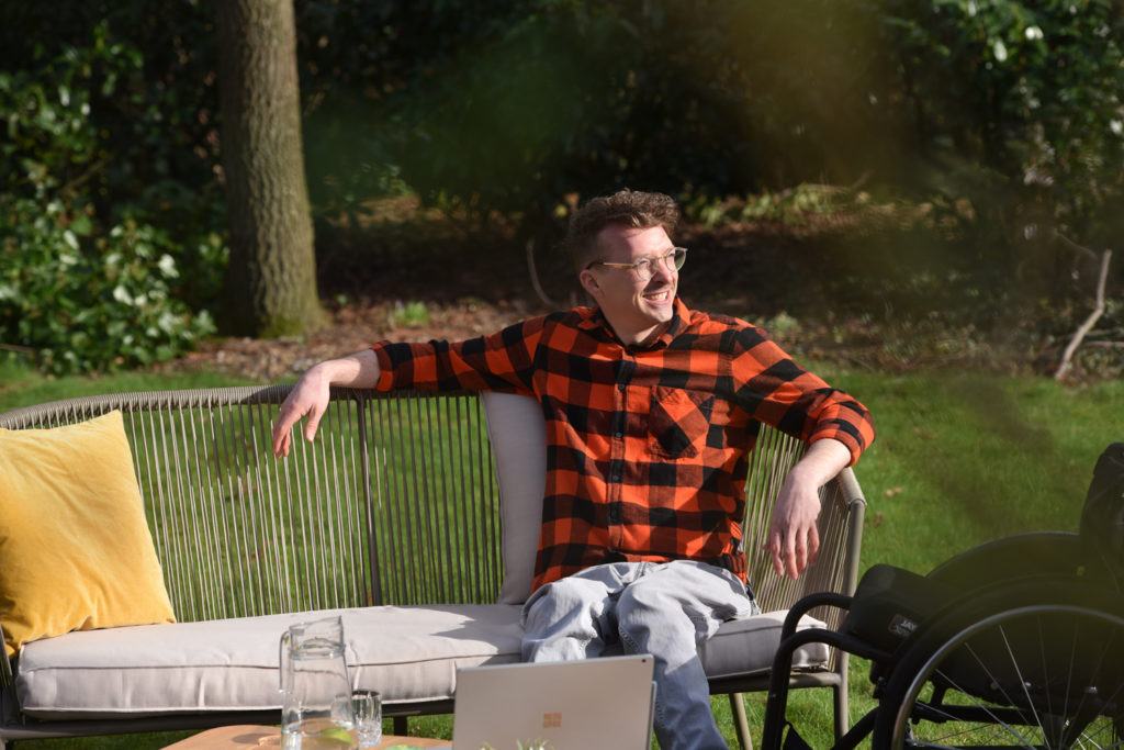 Dennis sitzt auf einer Bank in seinem Garten. Es ist ein sonniger Tag.