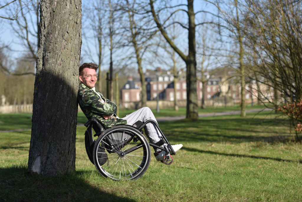 Dennis sitzt in seinem Rollstuhl und "kippelt" angelehnt an einem Baum. Es ist ein sonniger Frühjahrestag - die Bäume sind kahl.