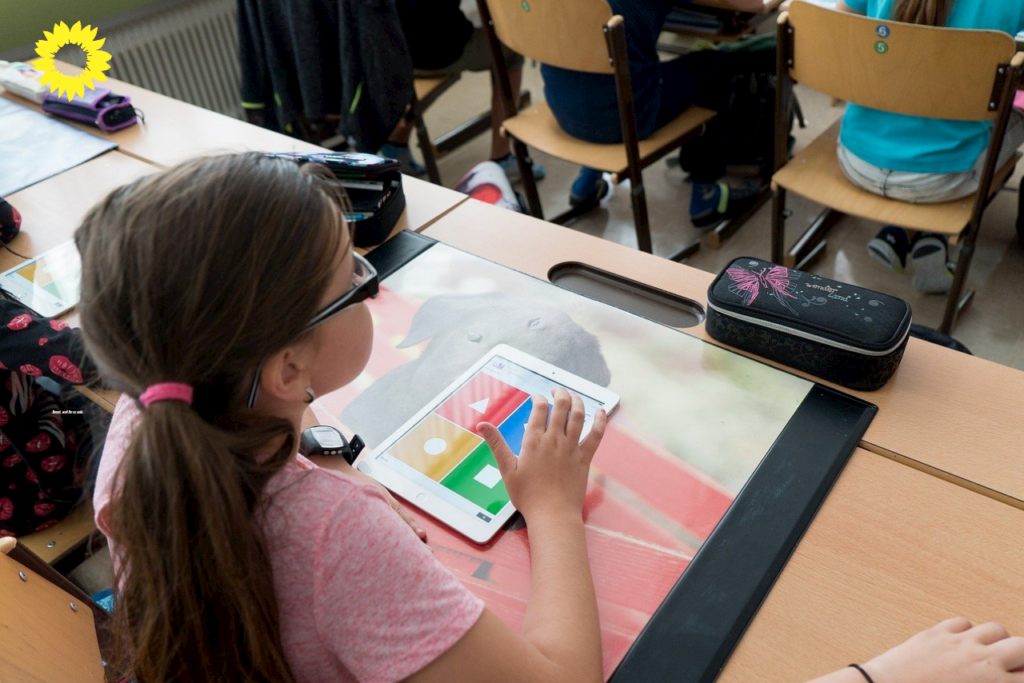 Auf dem Foto ist ein junges Mädchen mit einer Brille zu sehen. Das Mädchen sitzt am Tisch in der Schule und schaut nach vorne. Mit den Fingern tippt das Mädchen auf einem Tablet.