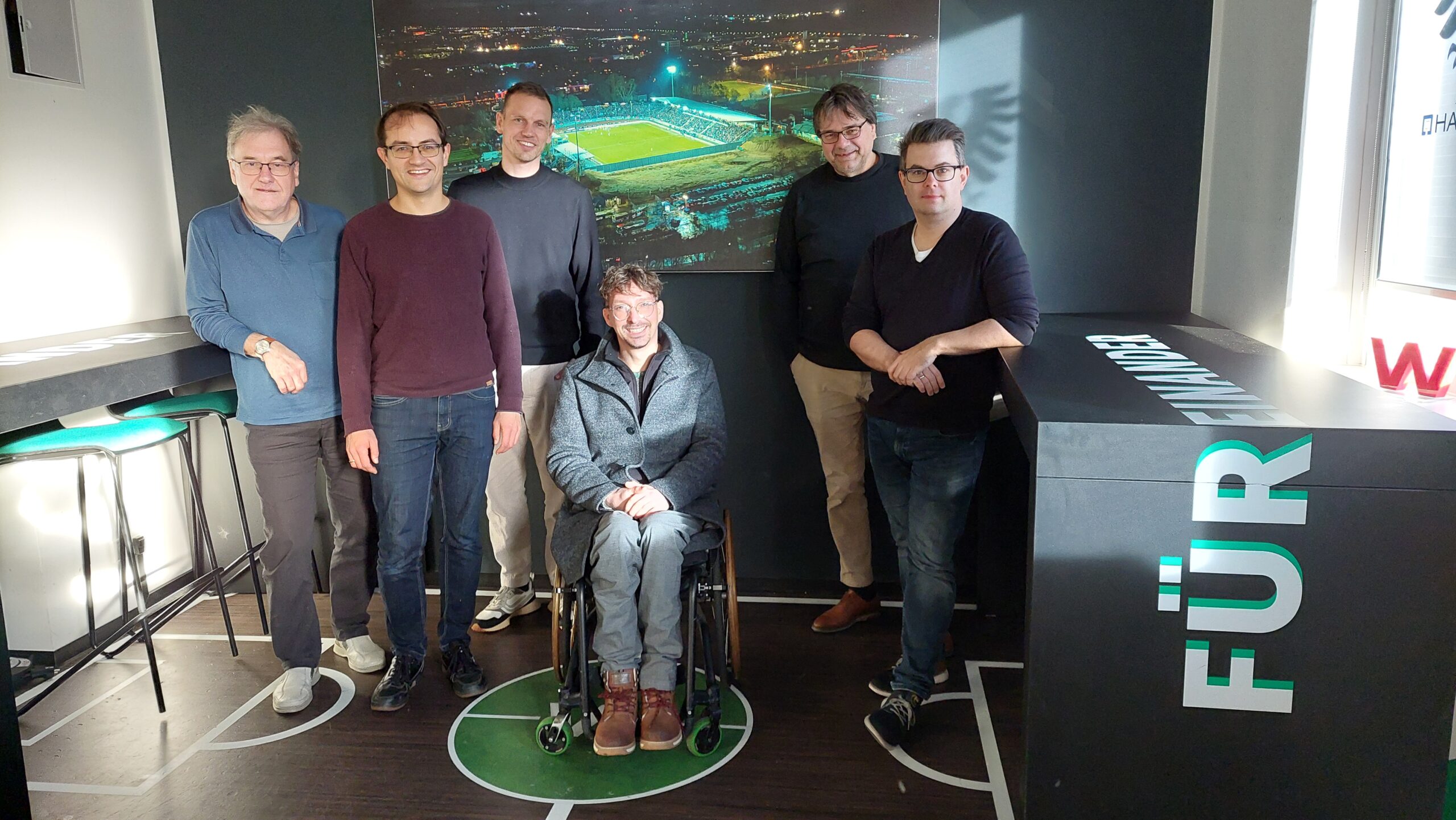 Eine Gruppe von Personen vor dem großen Bild des beleuchteten Stadion des SC Preußen Münster. Menschen von links nach rechts: Harald Wölter (Grüne Ratsfraktion Münster), Robin Korte MdL (GRÜNE), Dennis Sonne MdL (GRÜNE), Ole Kittner (Geschäftsführung SC Preußen Münster), Klaus Rosenau (Grüne Ratsfraktion Münster), Dr. Markus Sass (Geschäftsführung SC Preußen Münster)