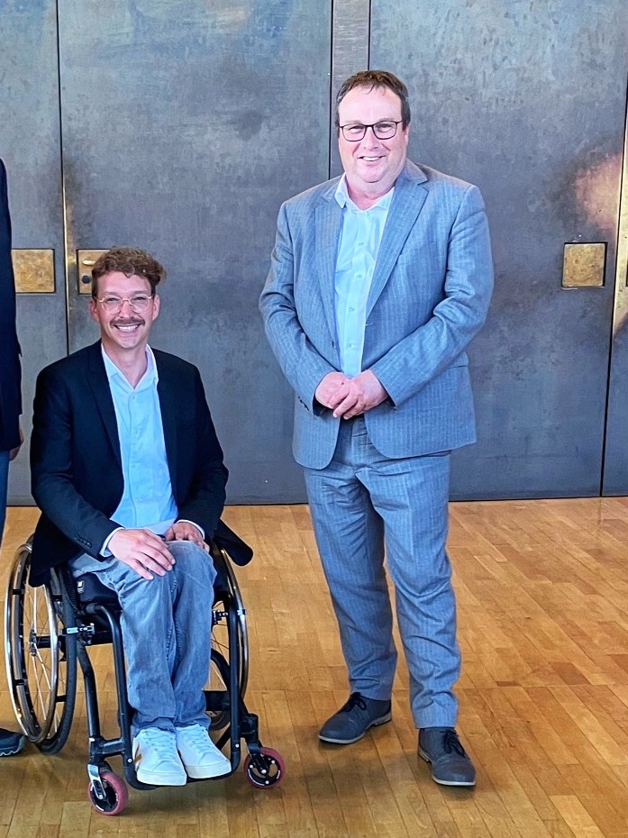Auf dem Bild sieht man Dennis Sonne im Rollstuhl sitzend und den NRW-Verkehrsminister Oliver Krischer.